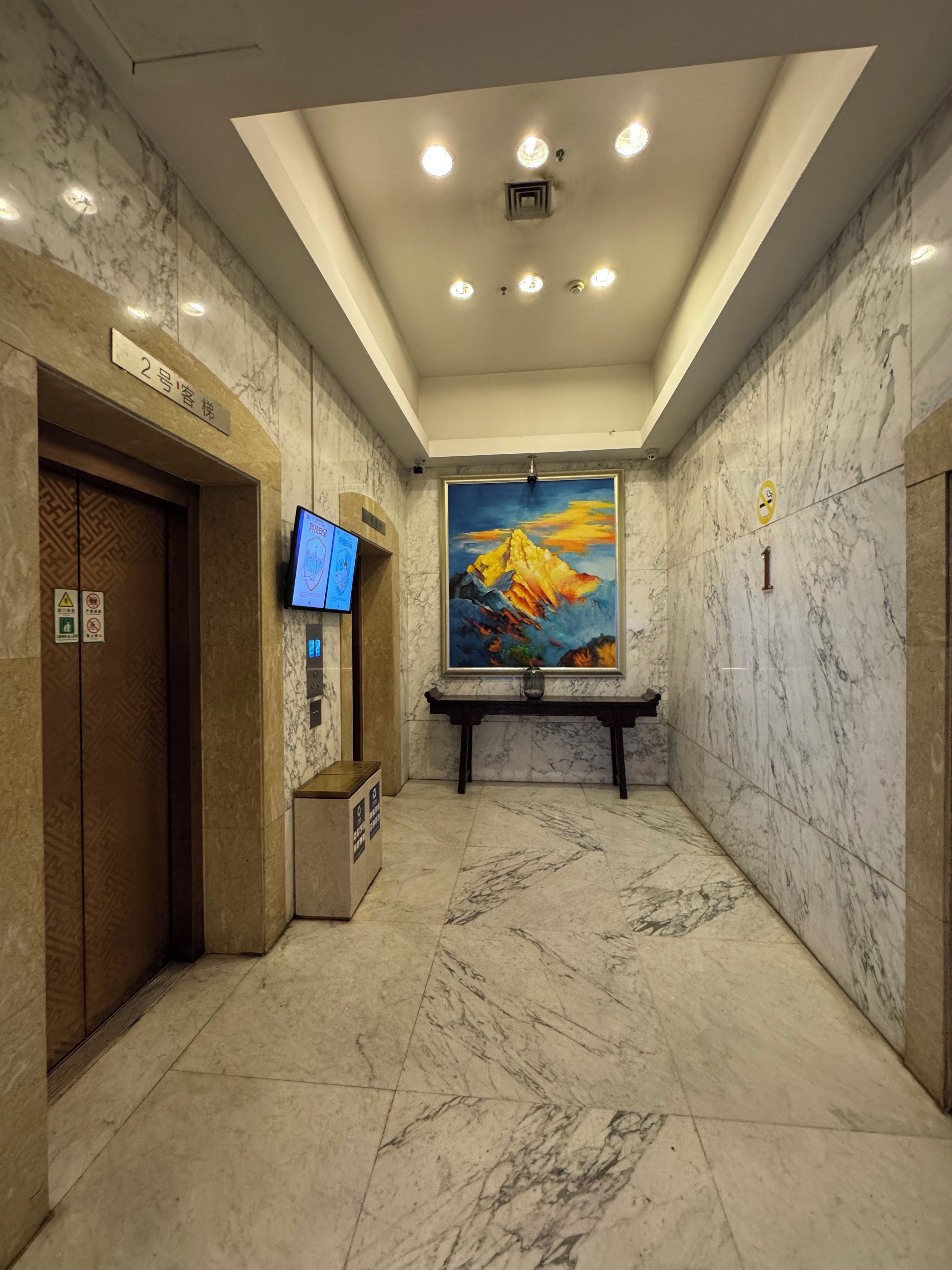 A lobby with marble floors, a digital information board, a painting, and an elevator.