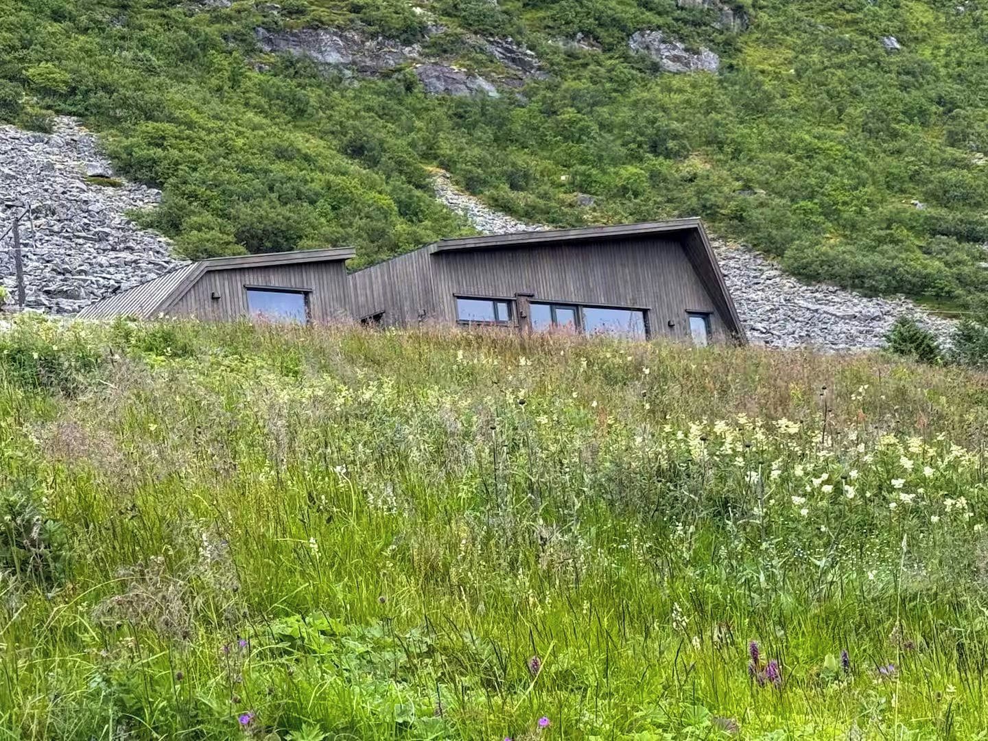 A modern building with a sloped roof, surrounded by lush greenery and rocky terrain.