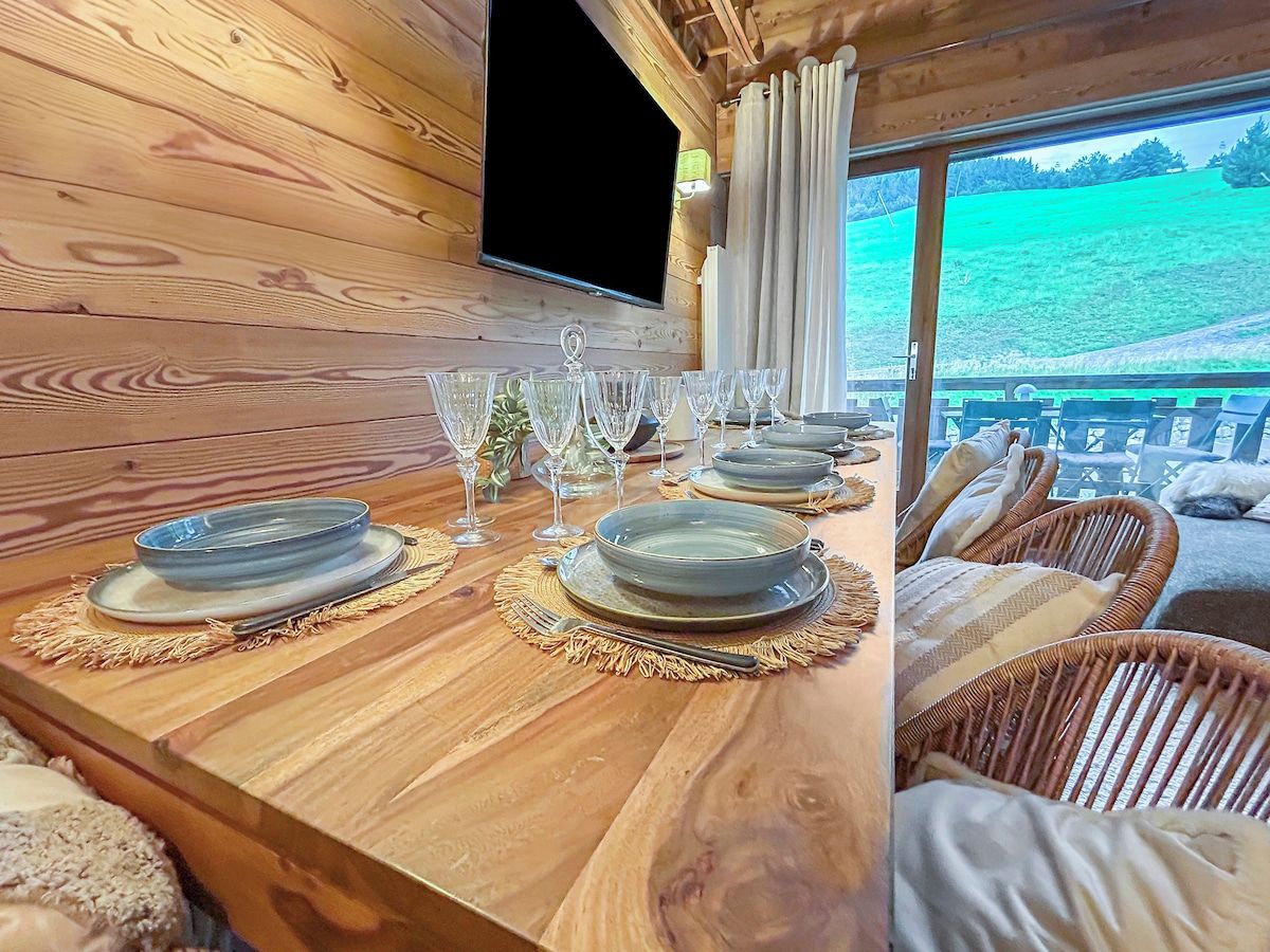 A wooden dining table set with plates, glasses, and cutlery, with a view of a green landscape through a window.