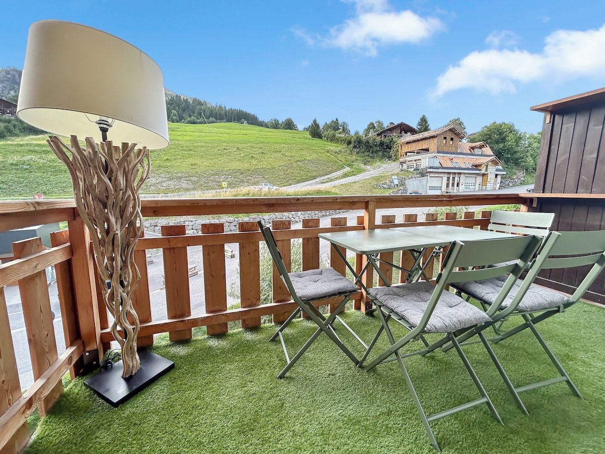 Een balkon met een tafel, stoelen, een lamp en uitzicht op een groen landschap en gebouwen.