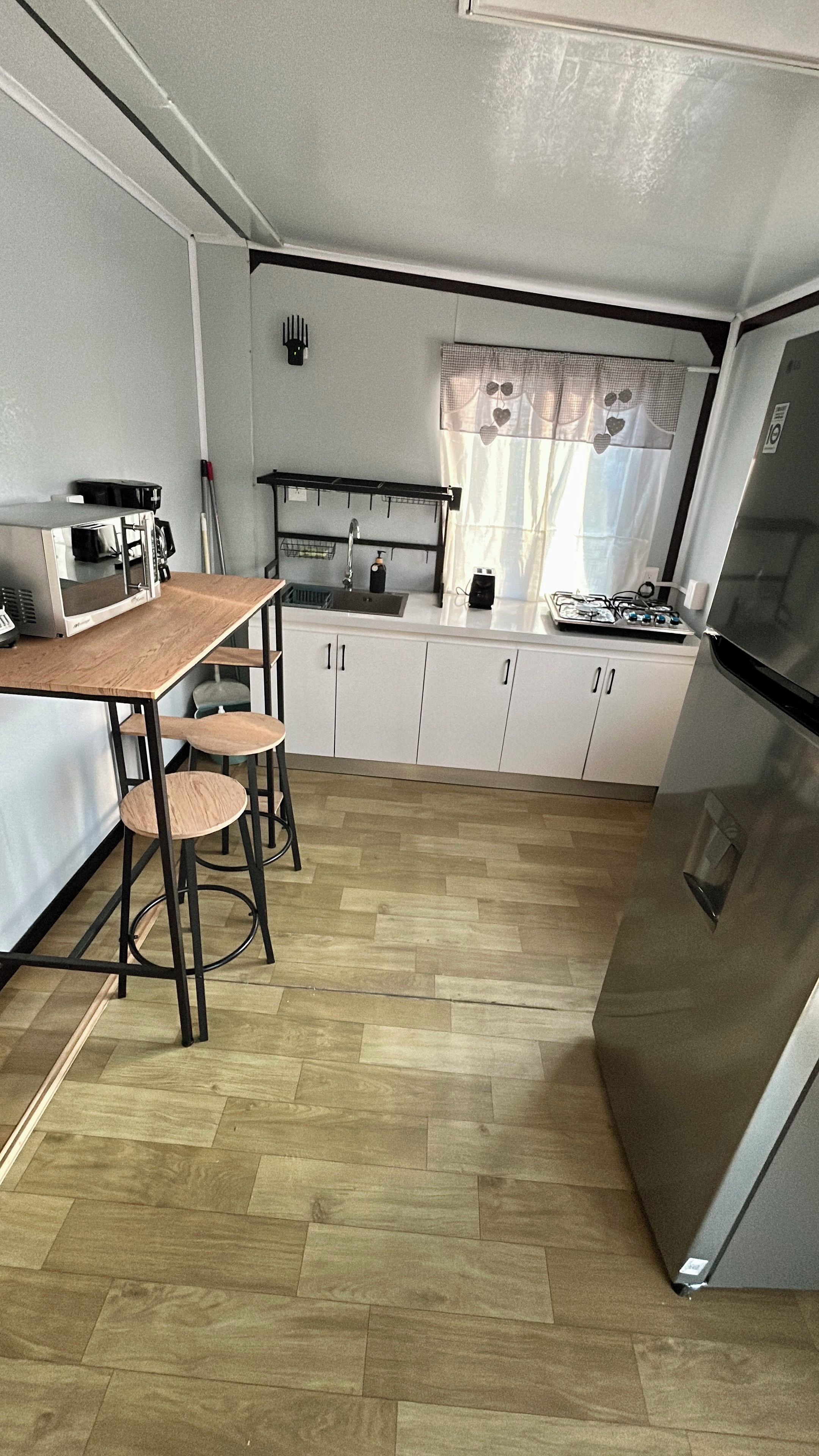 A compact kitchen with a wooden table, bar stools, a refrigerator, and a window with curtains.