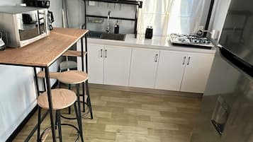 A compact kitchen with a wooden table, bar stools, a refrigerator, and a window with curtains.