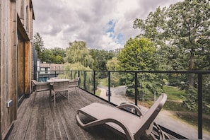 Una terraza de madera con mobiliario exterior, barandilla de vidrio y vistas a una carretera y árboles.