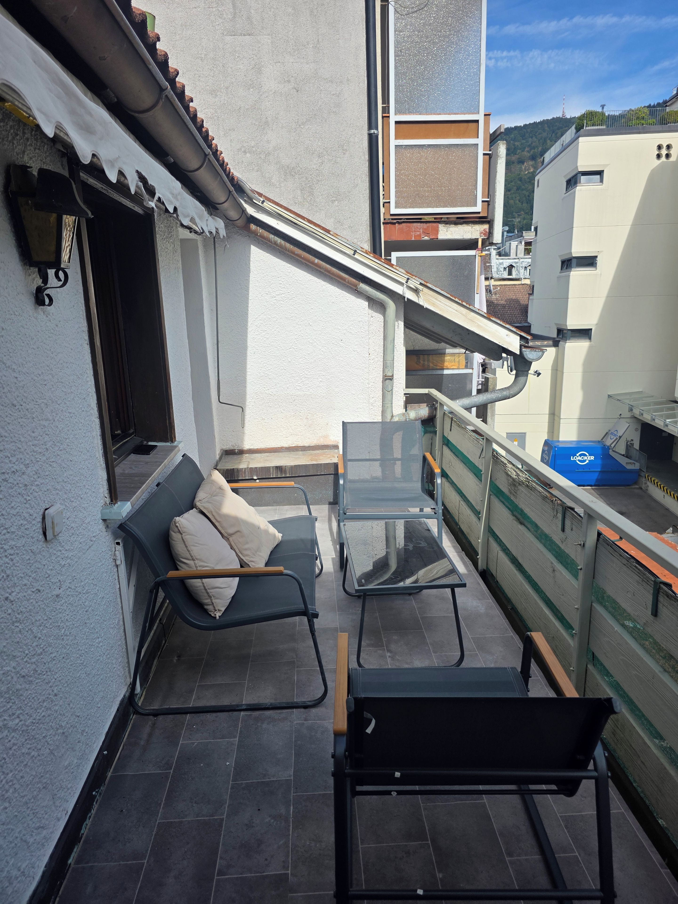 Ein Balkon mit einer Couch und zwei Stühlen, ein Fenster und ein Blick auf ein Gebäude im Hintergrund.