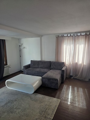 A living room with a sectional sofa, a white coffee table, a floor lamp, and sheer curtains. - E&H Appartements (Bregenz)