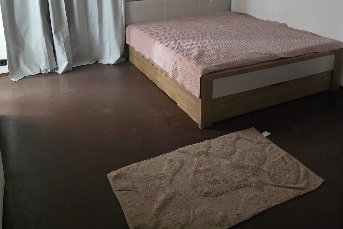 A bedroom with a bed, a window with curtains, a wall-mounted light, and a rug on the floor.