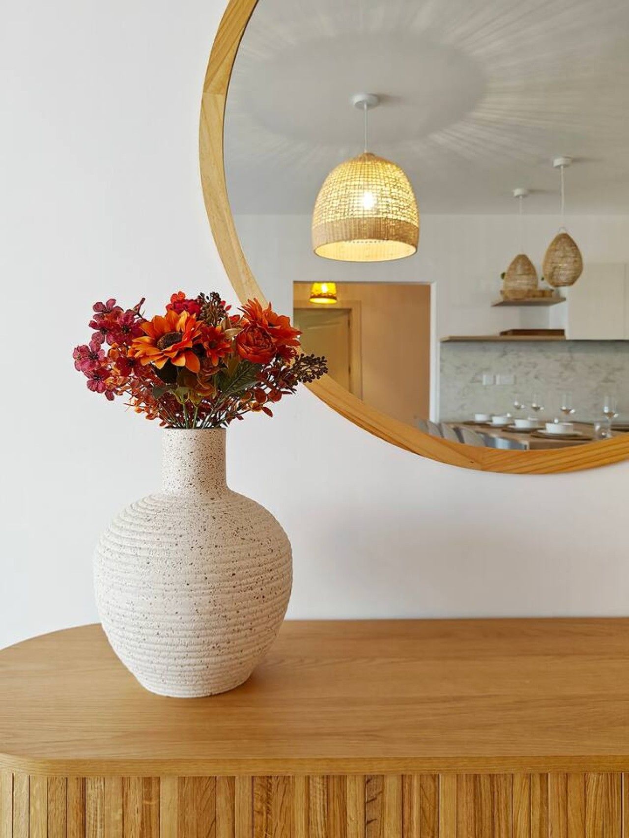 A round mirror reflecting a room with a wooden table, a vase with flowers, and a hanging light fixture.