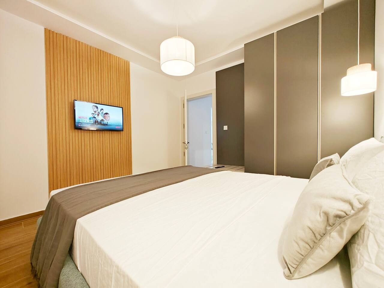 Une chambre d’hôtel moderne avec un grand lit, un téléviseur fixé sur un mur recouvert de panneaux en bois, et une porte donnant sur une autre pièce.