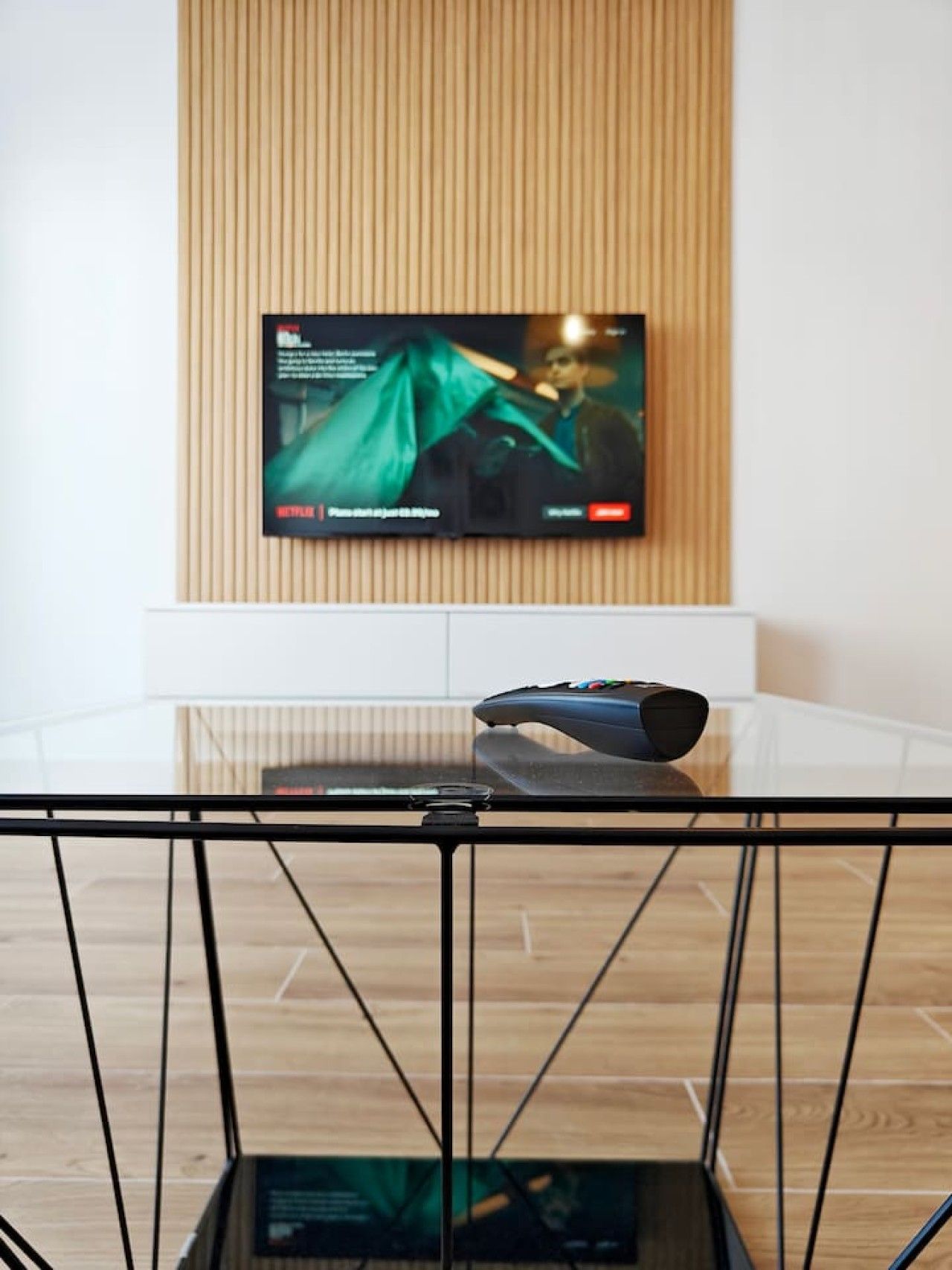 A television mounted on a wall with a wooden panel design, a remote control on a glass table, and a black metal chair in the foreground.