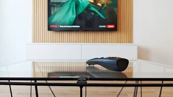 A television mounted on a wall with a wooden panel design, a remote control on a glass table, and a black metal chair in the foreground.