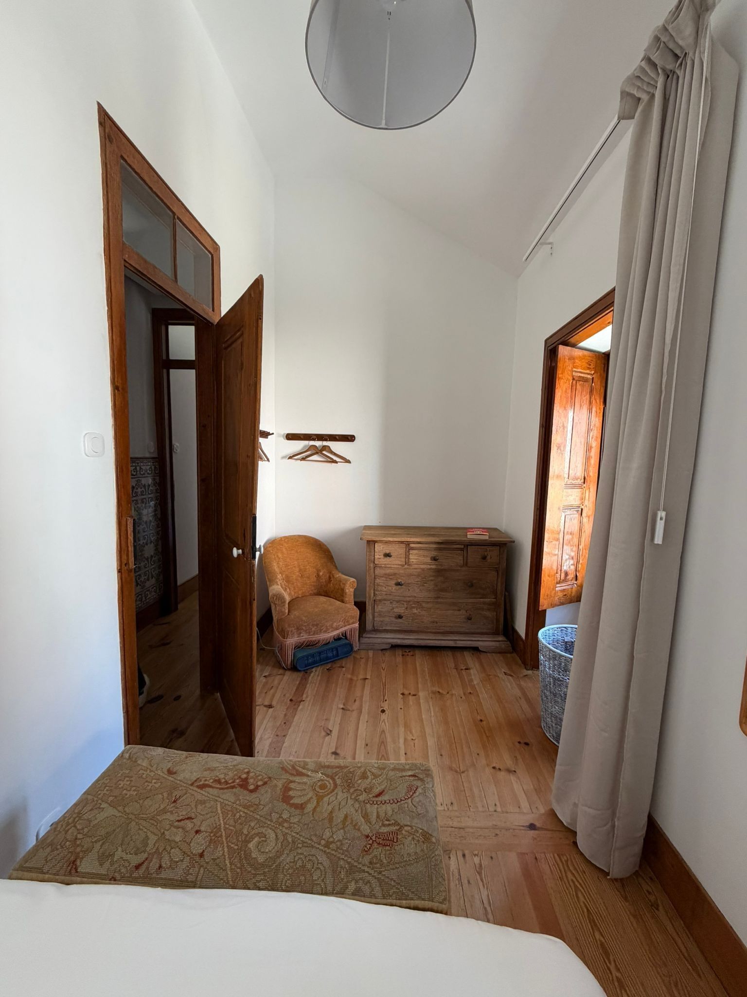 Un couloir étroit au plancher de bois, avec une commode en bois, une chaise et un lit recouvert d’une couvre-lit à motifs.