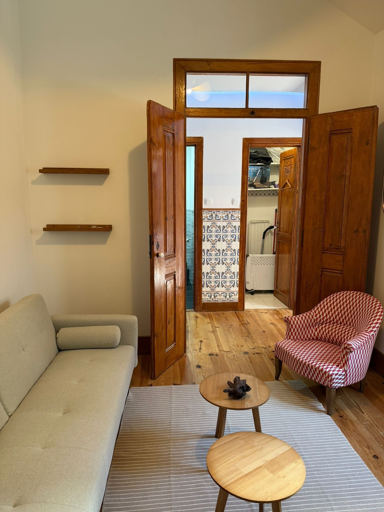 Una sala de estar con un sofá, un sillón estampado, una pequeña mesa de madera y vistas a un balcón con suelo de baldosas a través de una puerta abierta.