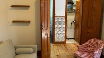 Una sala de estar con un sofá, un sillón estampado, una pequeña mesa de madera y vistas a un balcón con suelo de baldosas a través de una puerta abierta.