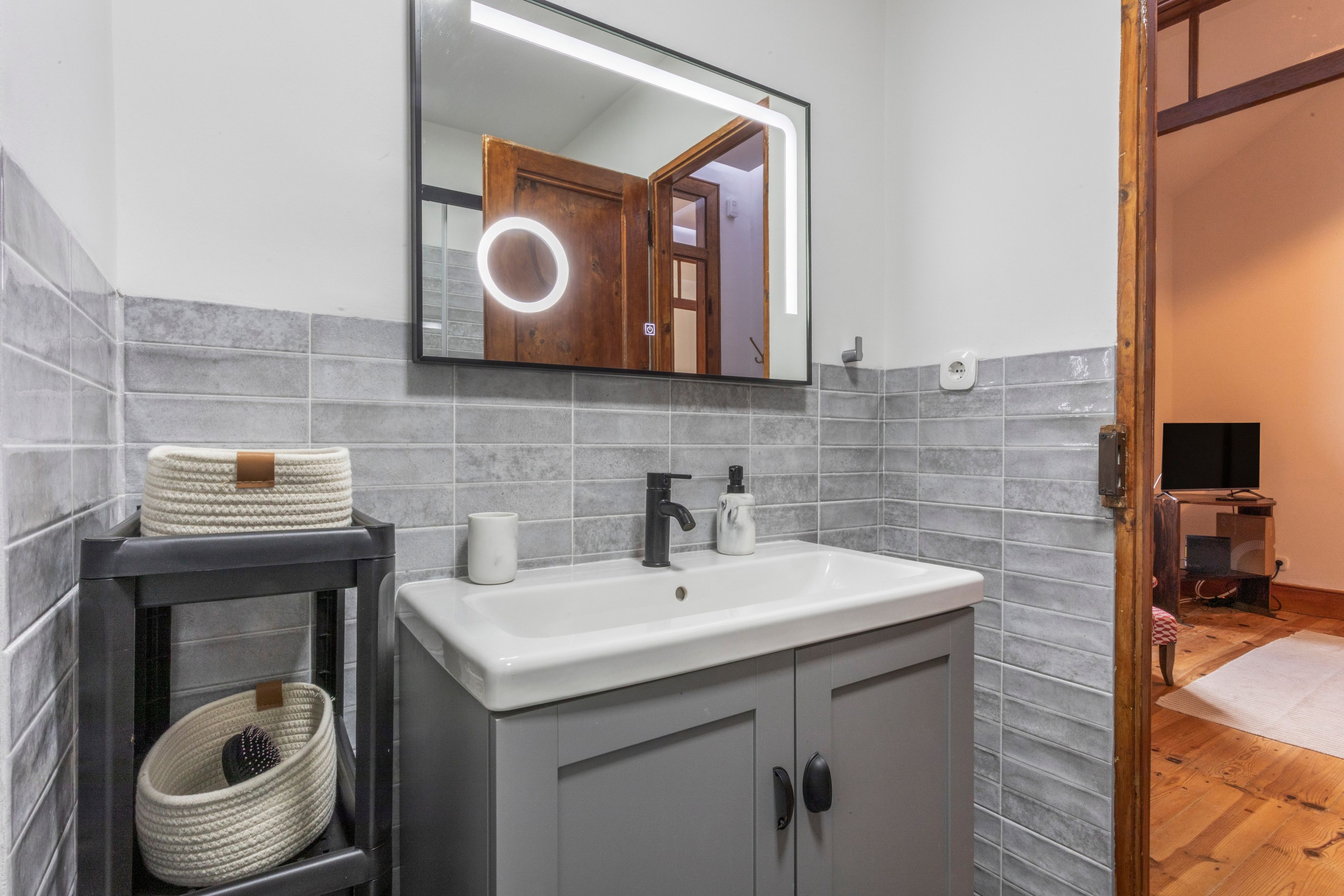 Une salle de bain avec un lavabo blanc, un miroir avec un éclairage intégré, des murs recouverts de carreaux gris, une porte en bois et une petite étagère sur laquelle se trouve un panier.