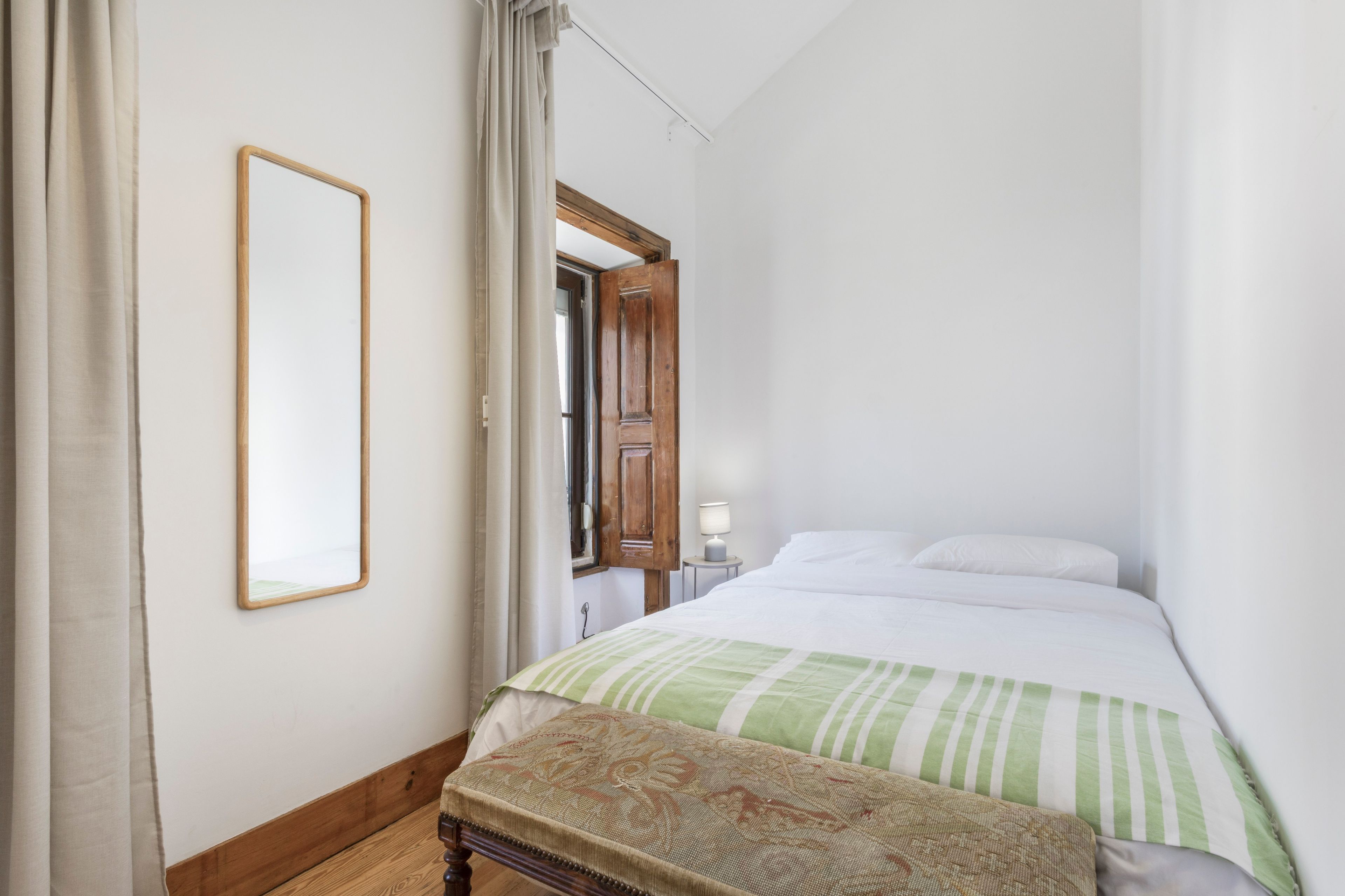 A bedroom with a bed, a bench, a bedside table with a lamp, and a mirror on the wall.
