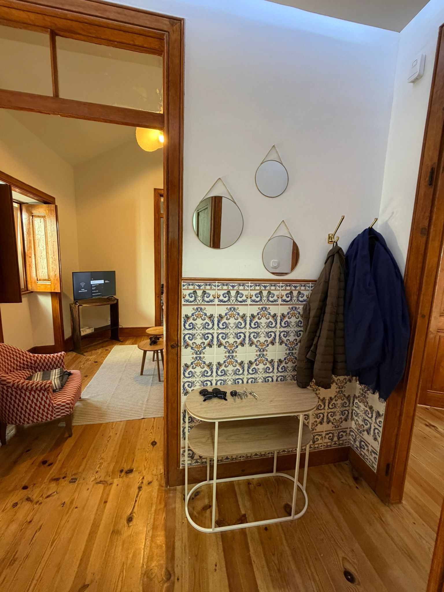 Una habitación con piso de madera, una mesita con estructura metálica, una silla, un perchero con una chaqueta y un televisor sobre un mueble.