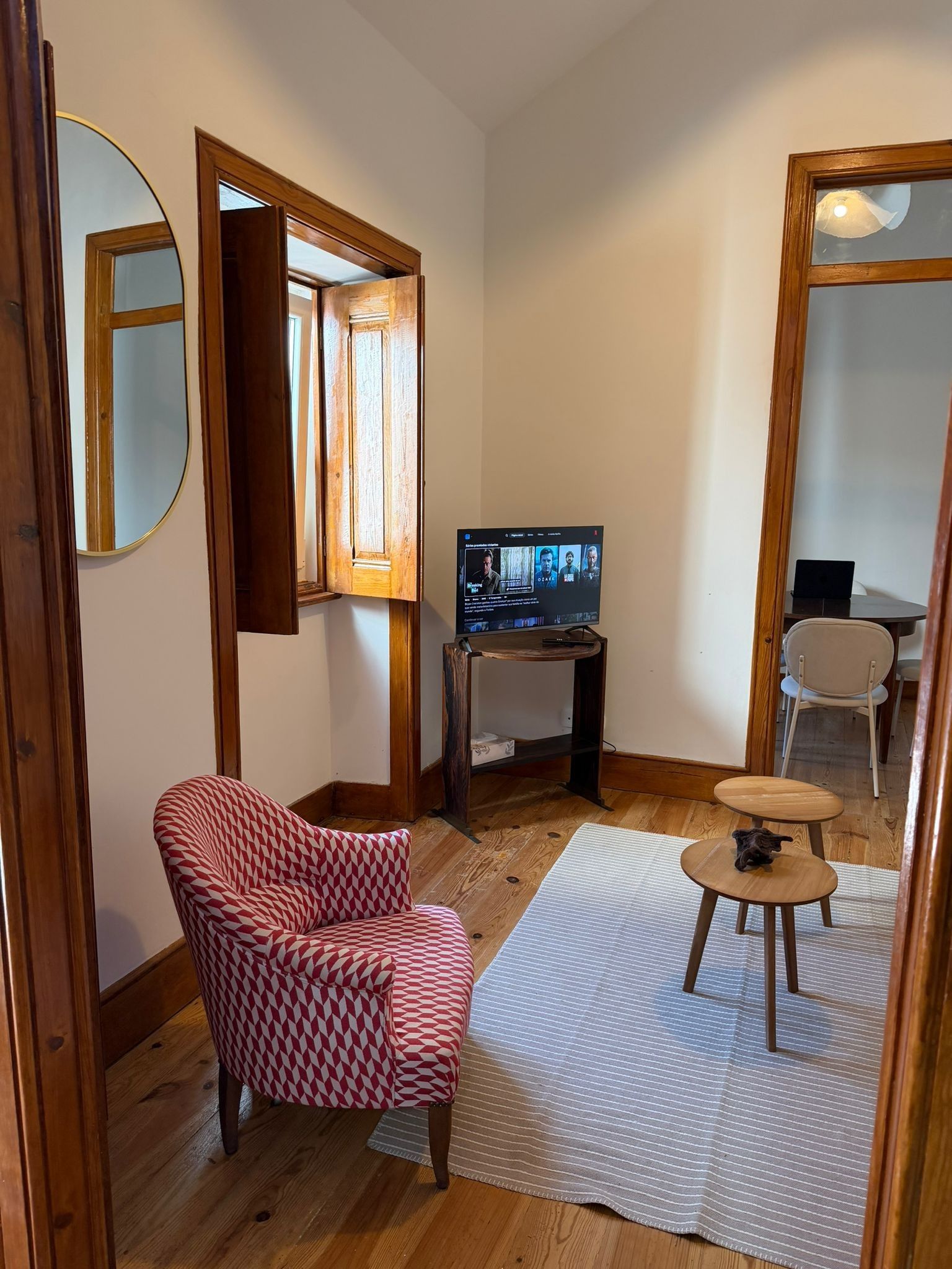 Une pièce chaleureuse avec un fauteuil à motifs rouges et blancs, une petite table ronde, un téléviseur posé sur un meuble en bois et un miroir accroché au mur.
