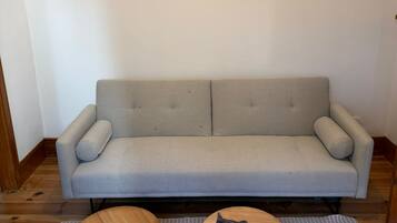 En sovesofa i neutrale farver med to puder, en stribet måtte og to runde træborde, hvor der ligger en mørk genstand på det ene.