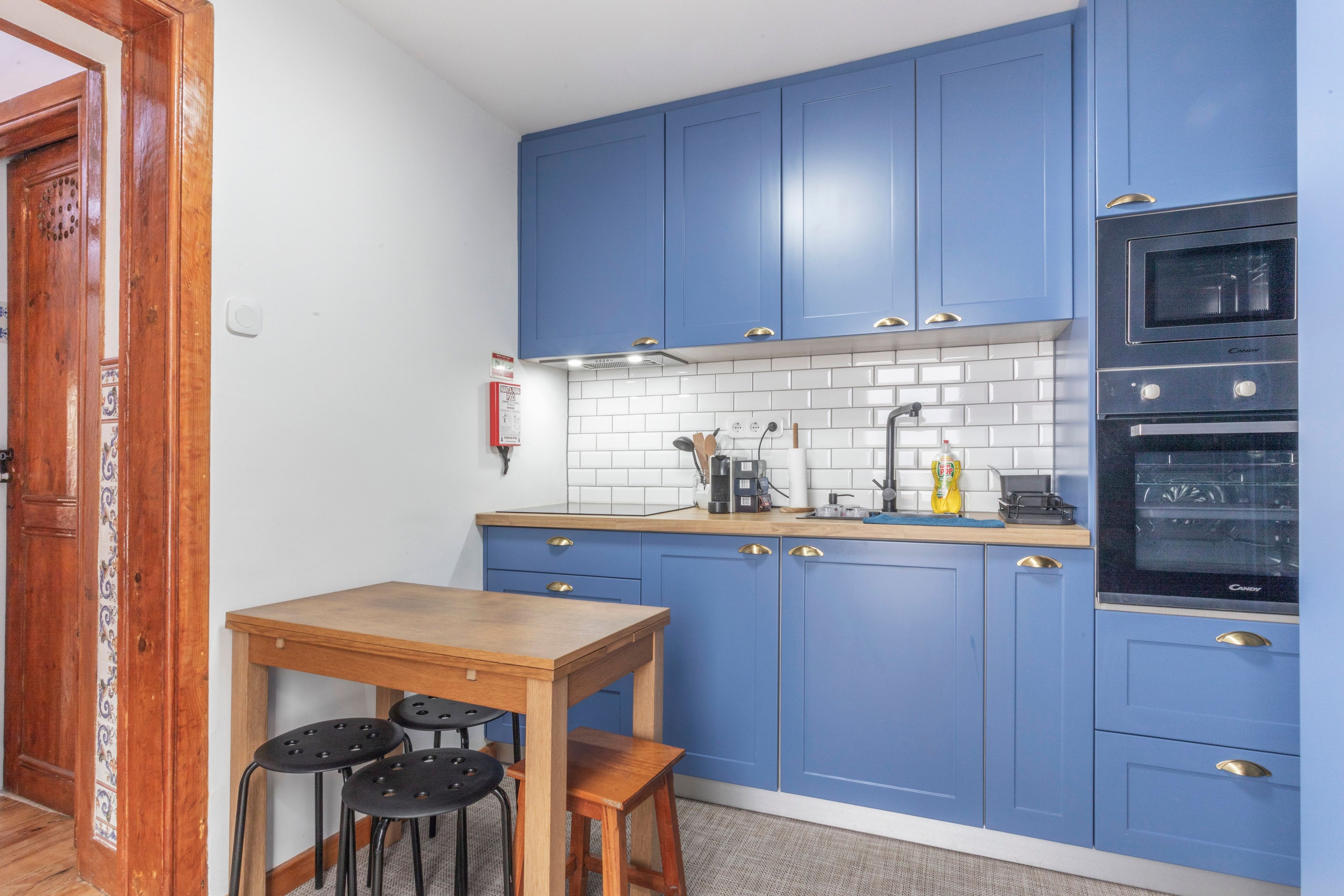 A compact kitchen with blue cabinets, a wooden table, and two stools.