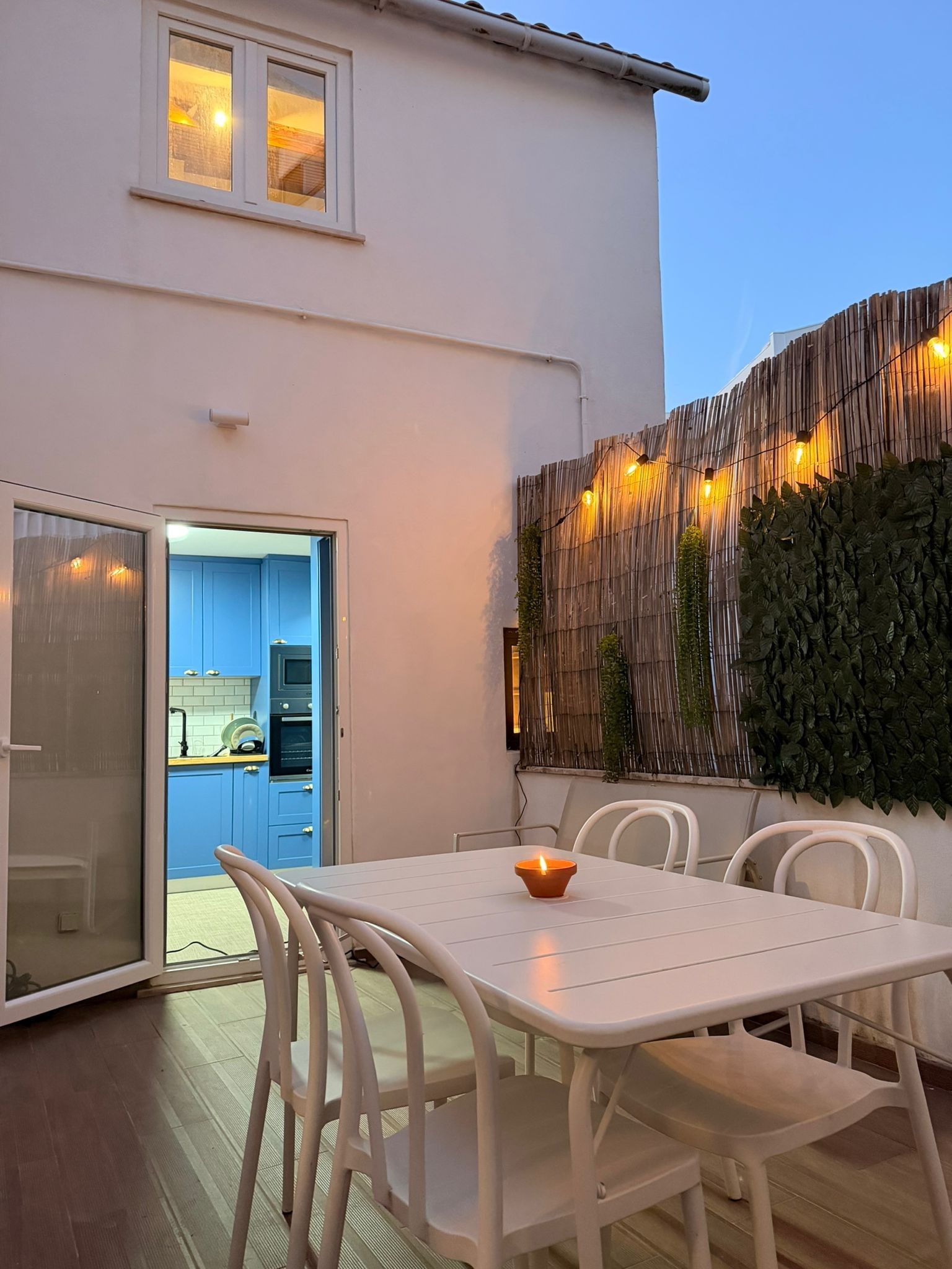 Terrassa exterior amb taula i cadires blanques, una espelma petita encésa i vista d'un espai interior blau.
