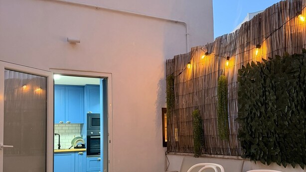 Eine Terrasse im Freien mit einem weißen Tisch und Stühlen, einer kleinen, beleuchteten Kerze und Blick auf einen blauen Innenraum.
