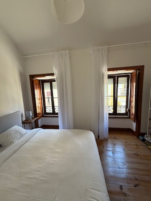 Un dormitori amb un llit gran, dues finestres amb cortines, un paviment de fusta i una petita taula de nit.