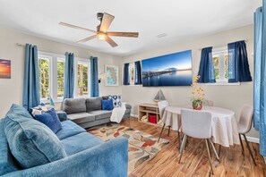 Ein Wohnzimmer mit einer Couch, einem Esstisch mit Stühlen, einem Deckenventilator und einem großen Fenster mit Vorhängen.