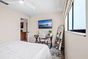 Ein Hotelzimmer mit einem Bett, einem Schreibtisch, einem Stuhl, einer Schreibtischlampe, einem Blumentopf, einem großen Spiegel und einem Fenster.