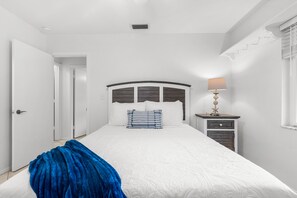 A bedroom with a bed, a bedside table with a lamp, and a blue throw blanket on the floor. - 212 Surf Road (Melbourne Beach)