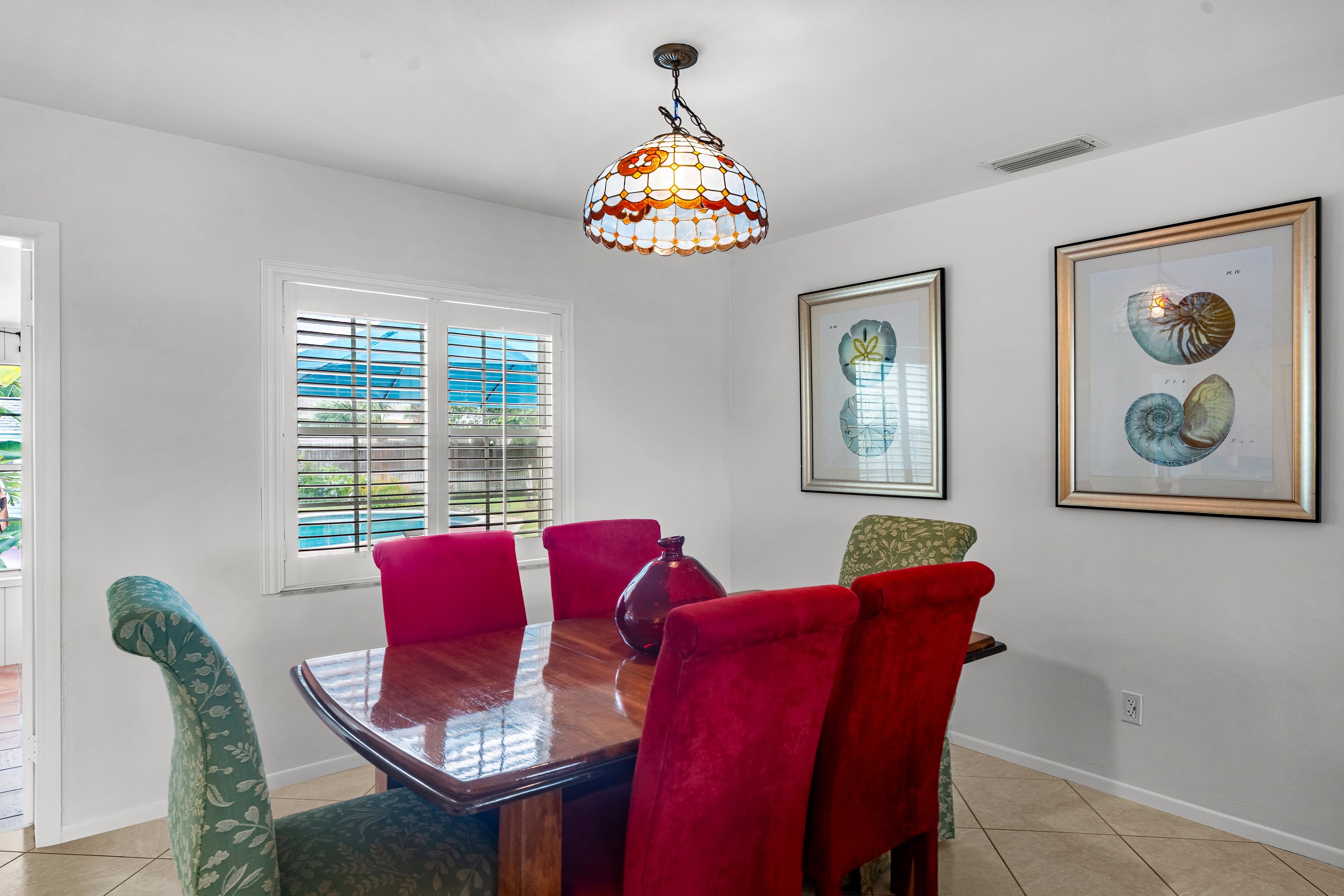 A dining area with a wooden table, red upholstered chairs, a stained glass pendant light, and framed artwork on the wall.