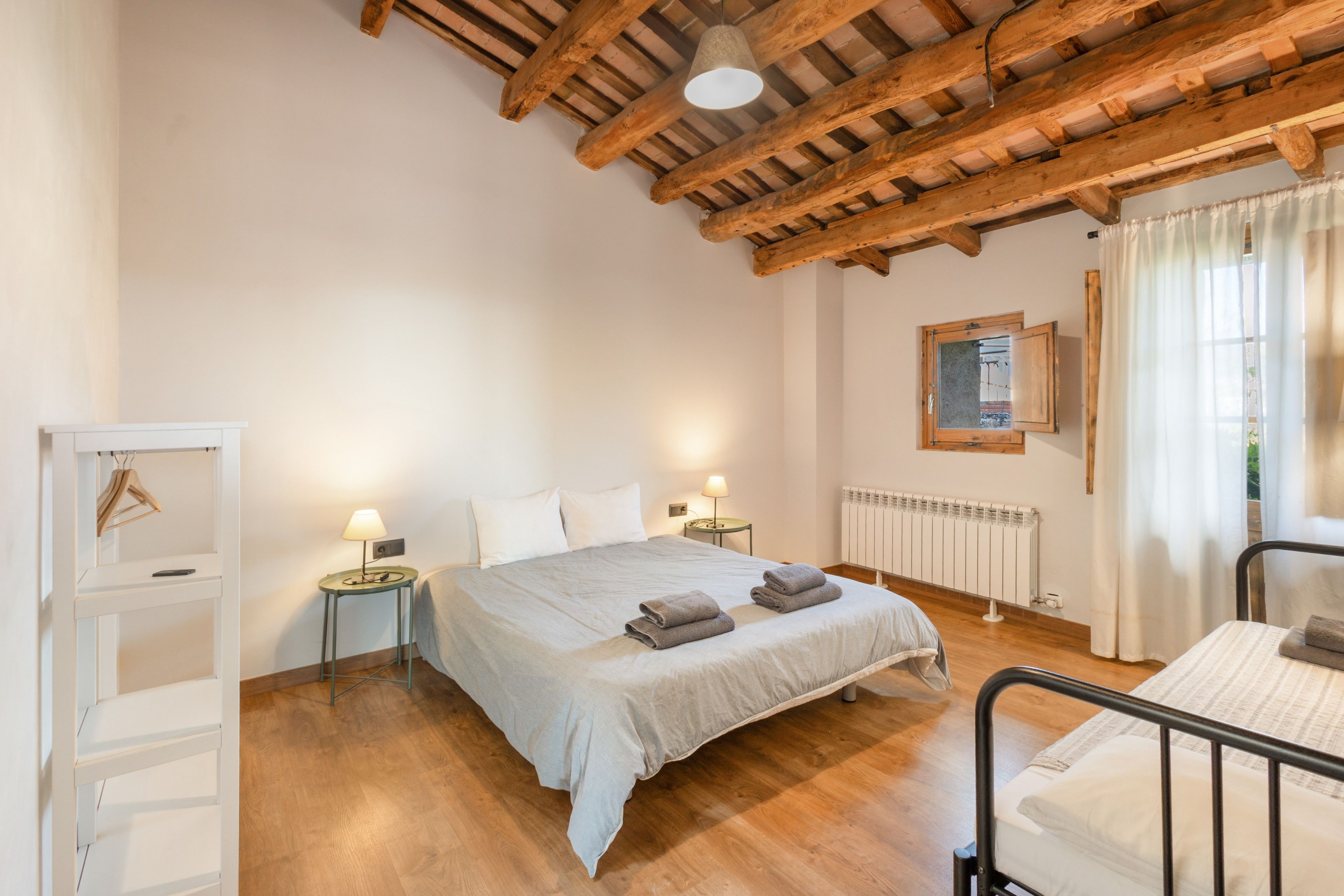 Une chambre avec un plafond en bois, un lit avec des oreillers et des serviettes, des tables de chevet avec des lampes, un radiateur et une fenêtre avec des rideaux.