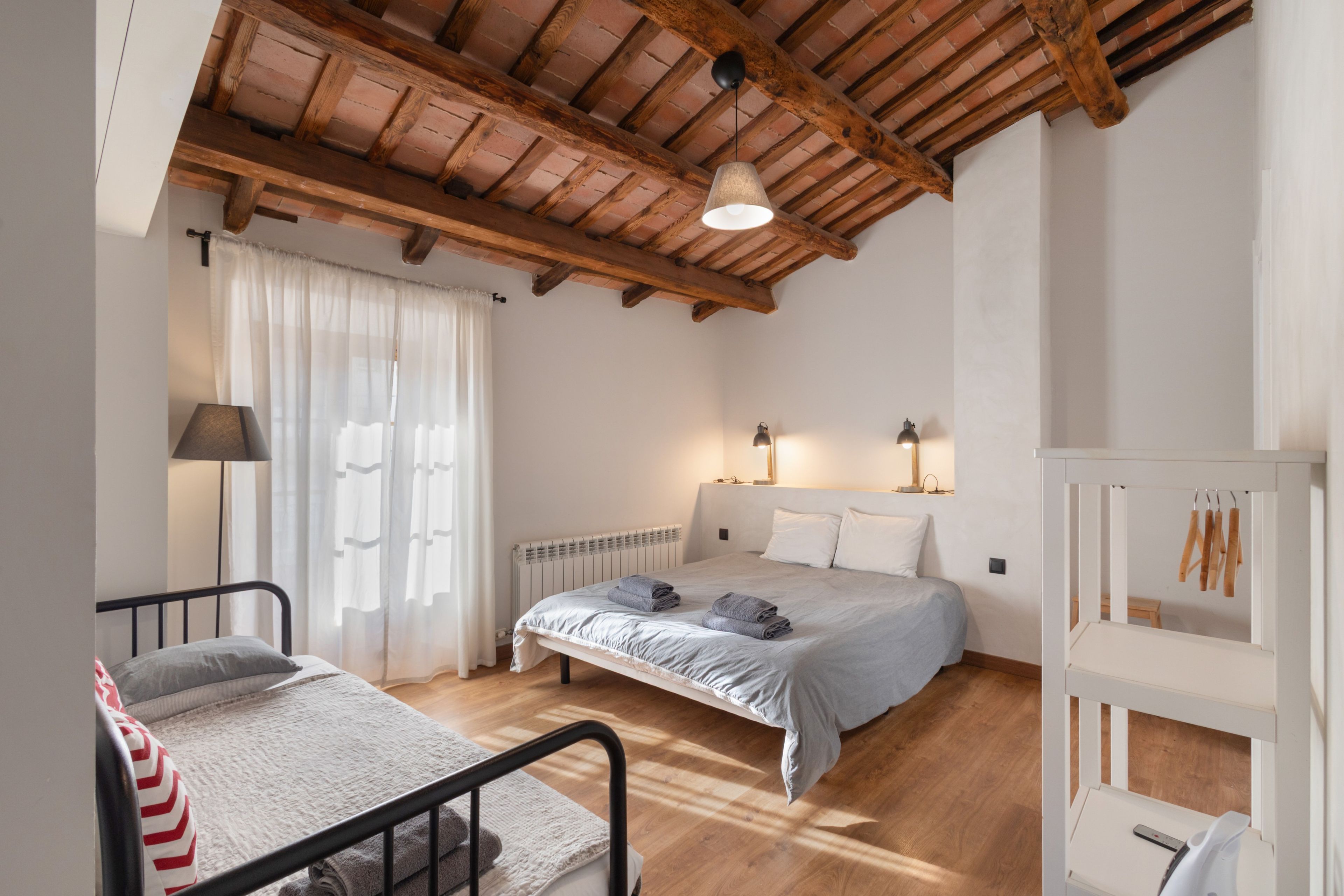 Un dormitorio con cama, lámparas de noche, un radiador, una ventana con cortinas y un techo de madera.