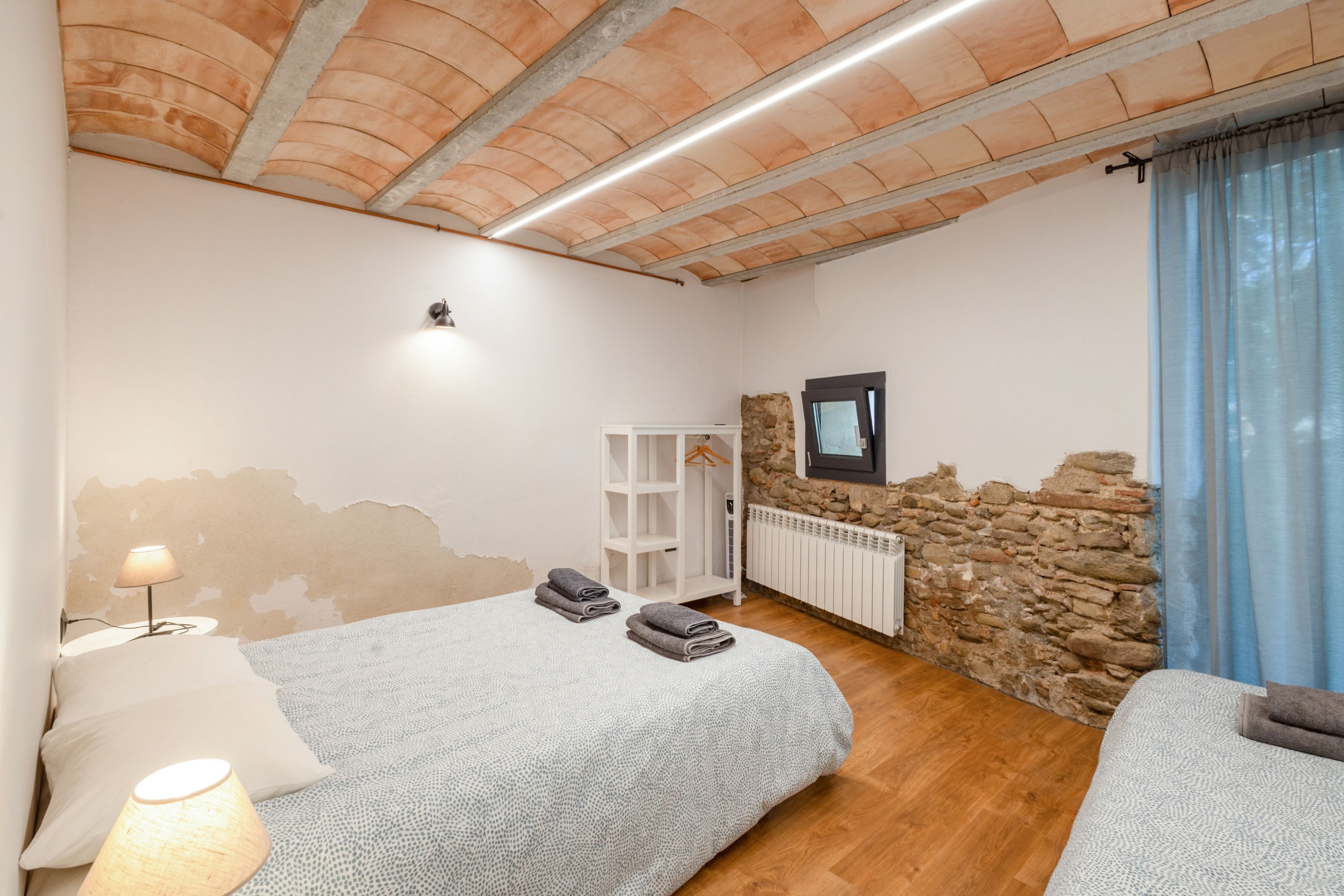 Un dormitorio con pared de piedra, una cama con colcha blanca texturizada, una mesita de noche con lámpara y un armario.