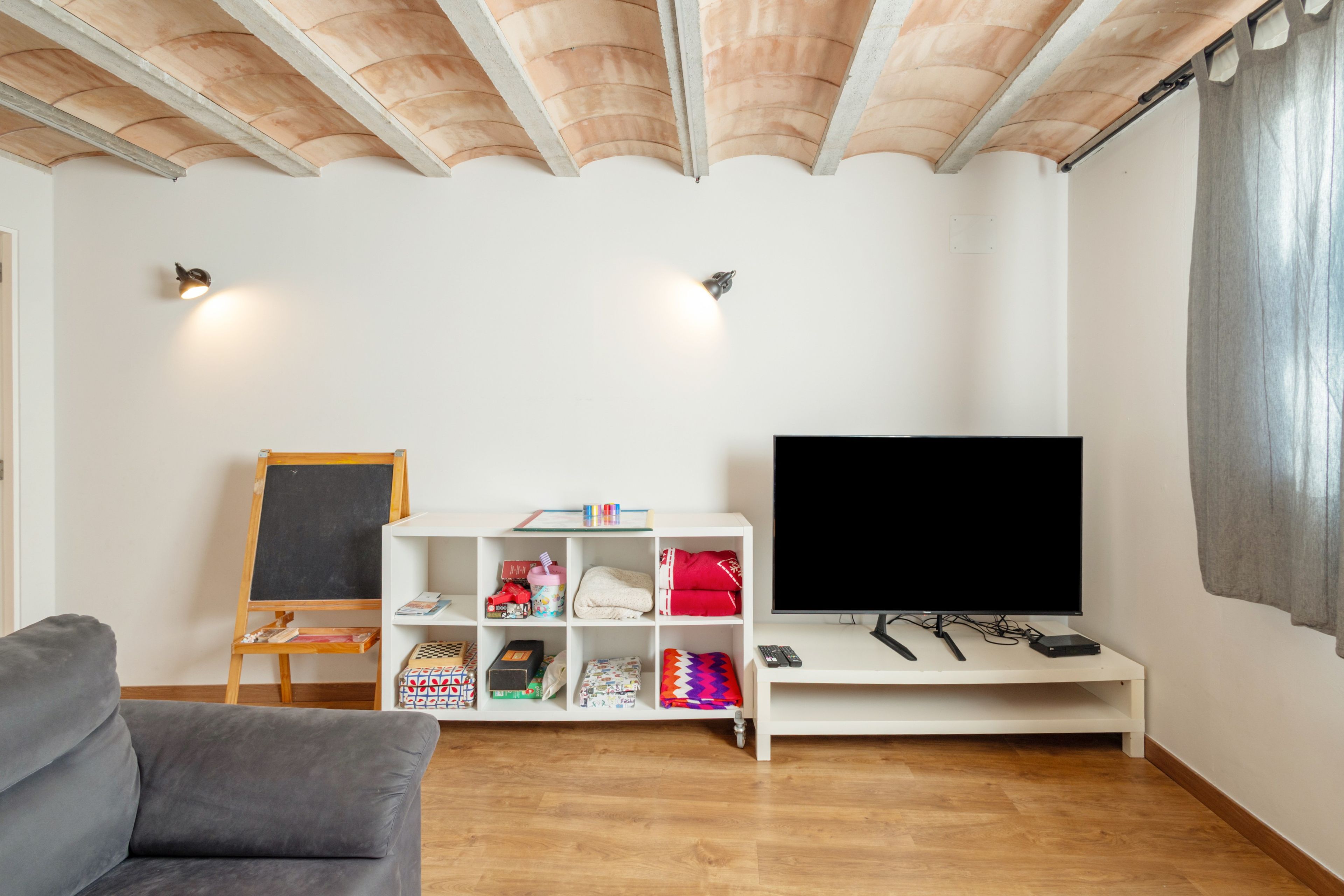 Una sala de estar con una televisión de pantalla plana, un mueble blanco con juguetes infantiles, una pizarra y un sofá gris.