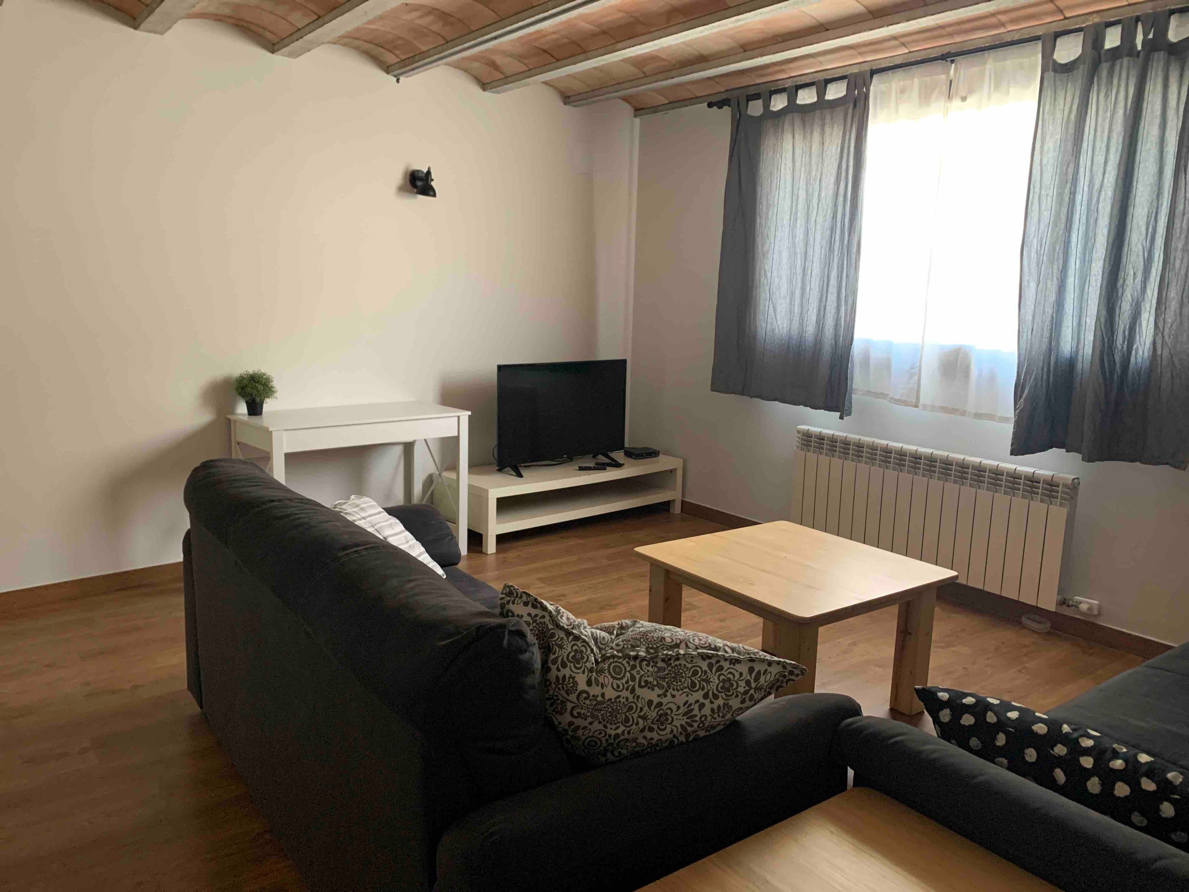 Una sala de estar con sofá, una mesa de centro, televisor sobre un mueble bajo y una ventana con cortinas.