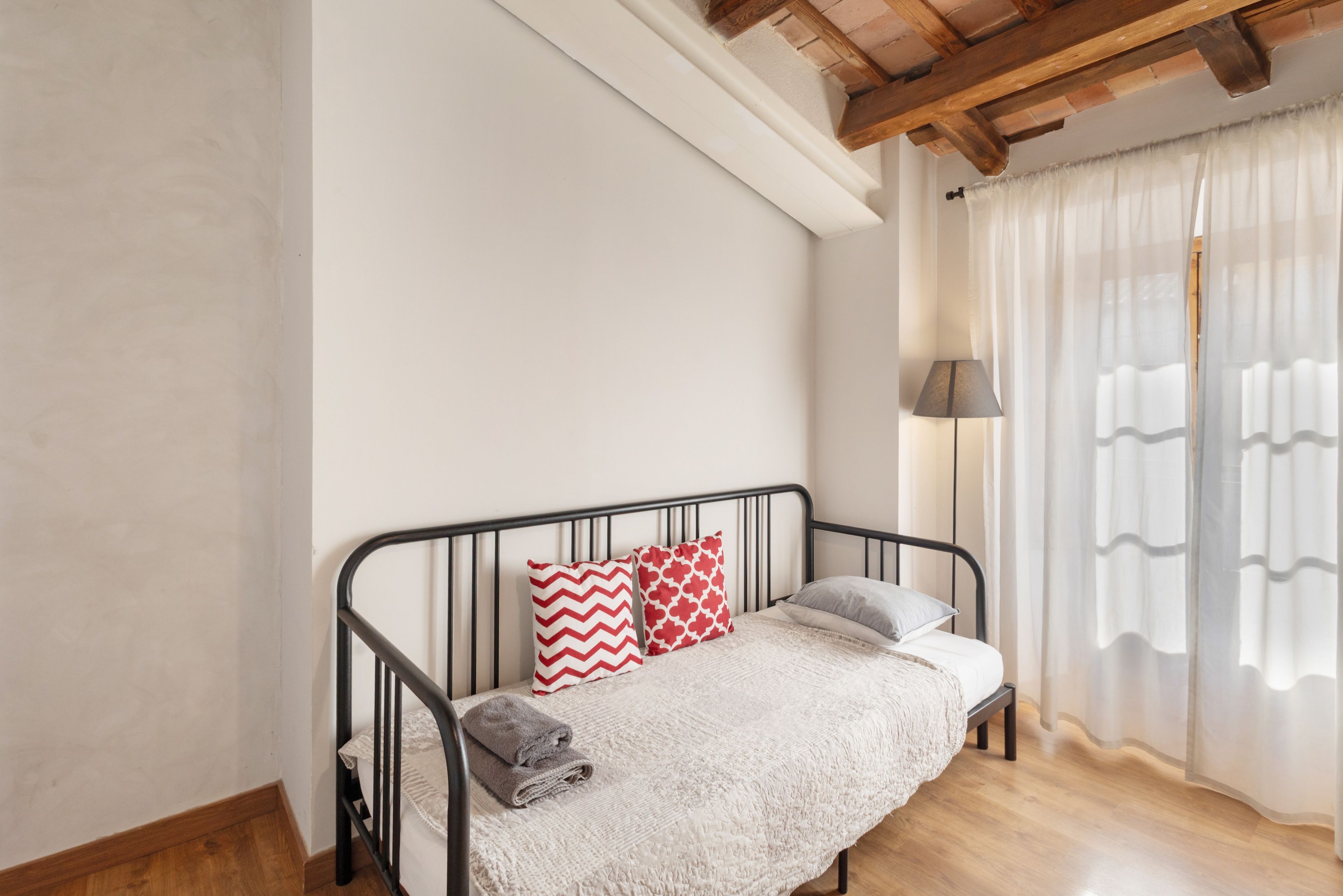 A single bed with a headboard, a bedside lamp, and a window with sheer curtains.