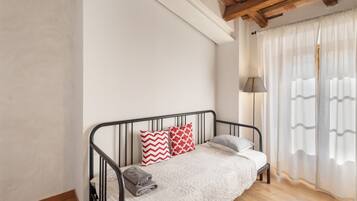 A single bed with a headboard, a bedside lamp, and a window with sheer curtains.