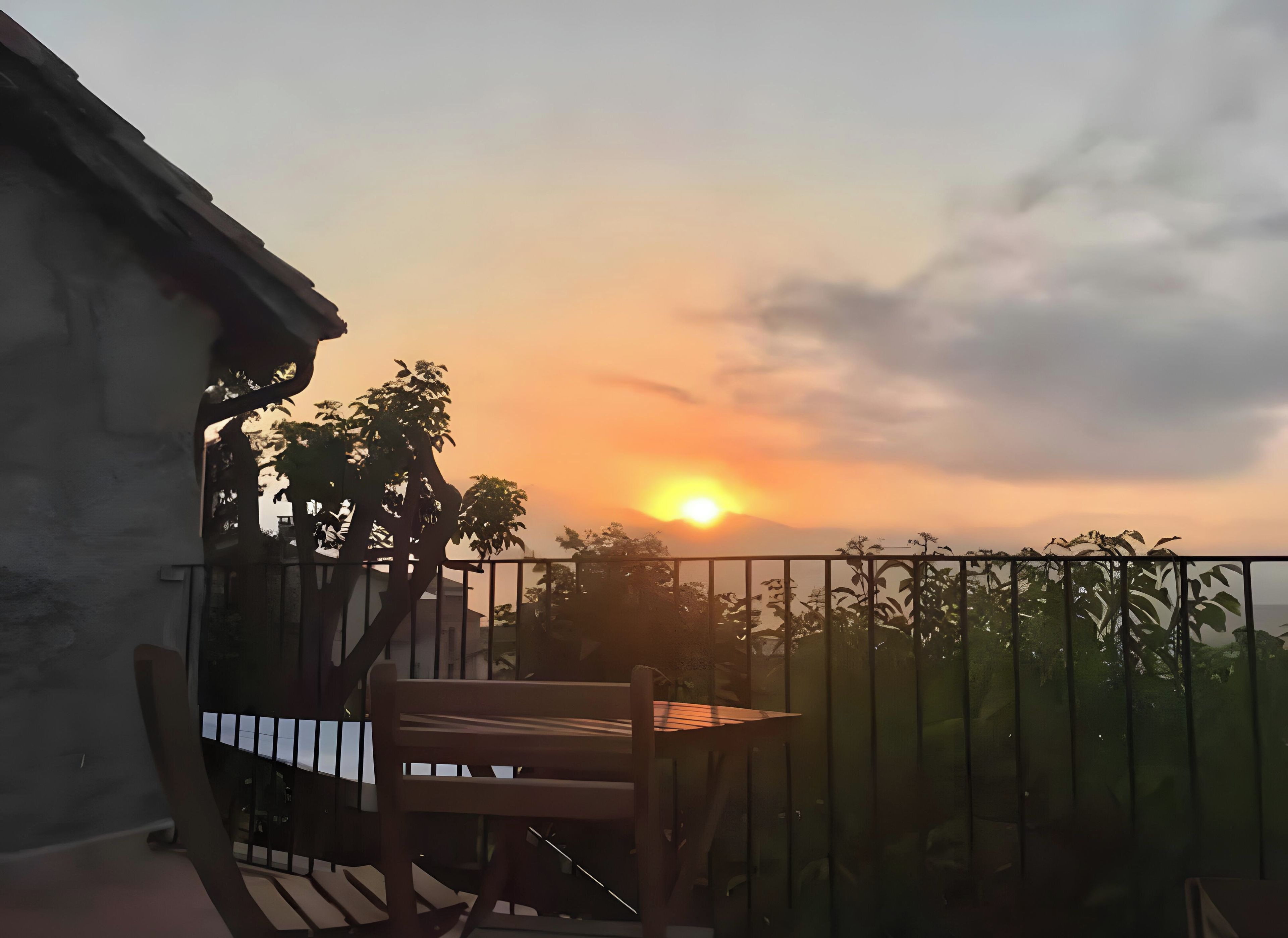 Un balcón con un banco y una mesa de madera, con vista a una puesta de sol y el sol bajo en el horizonte.