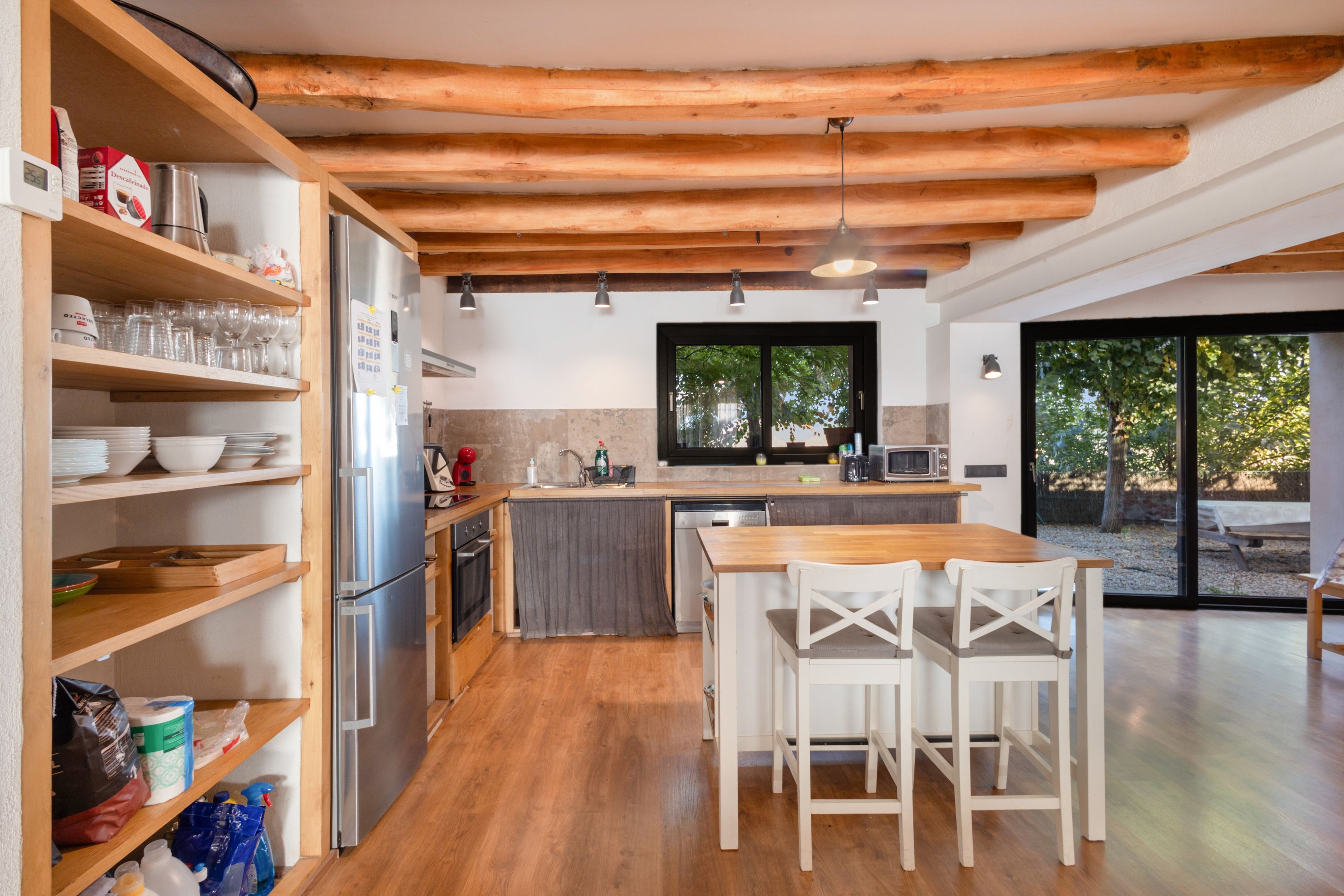 Cocina moderna con vigas de madera, refrigerador empotrado y una zona de comedor con sillas blancas.