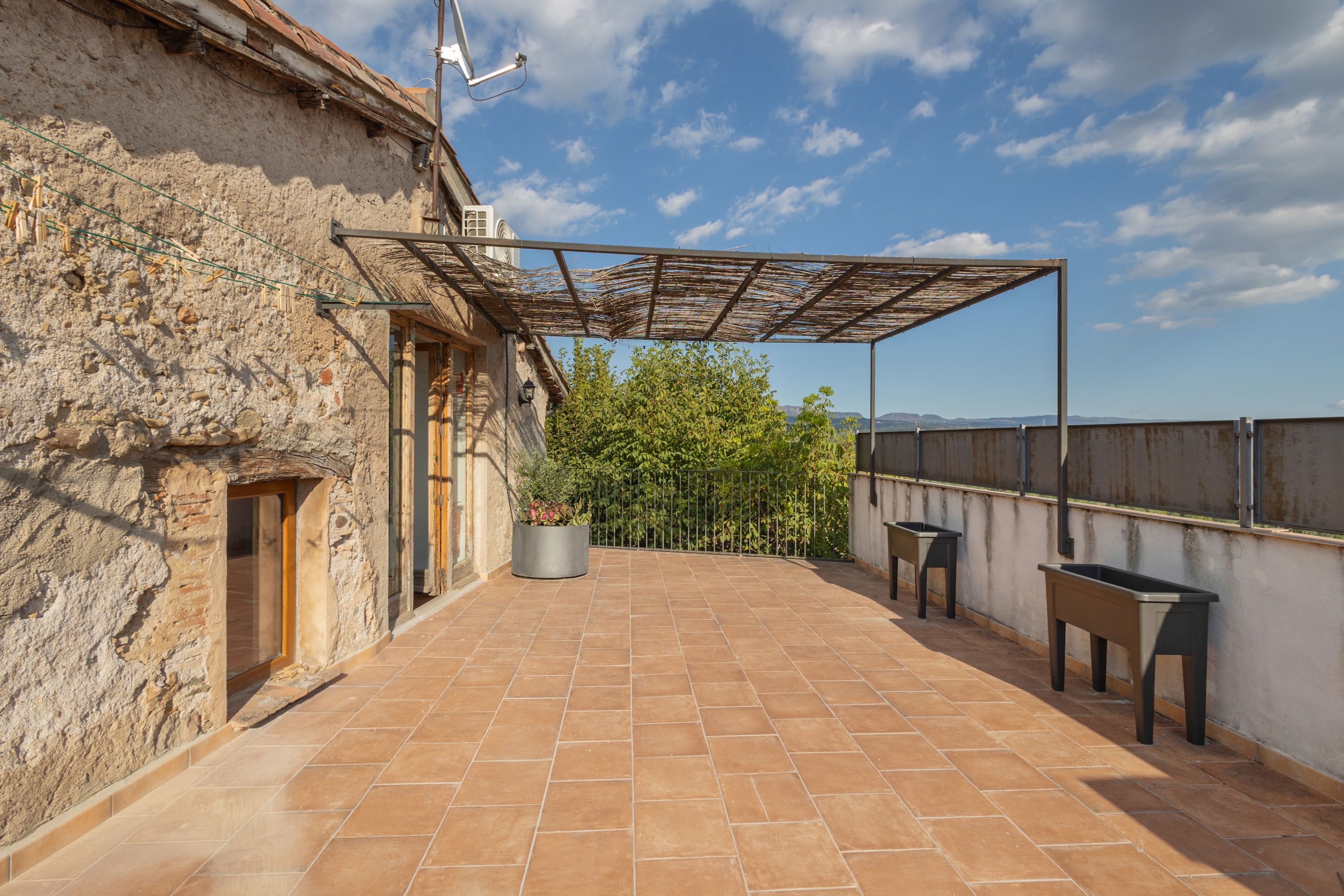 Terraza en la azotea con tejas de terracota, una pérgola, una planta en maceta y dos mesas de metal.