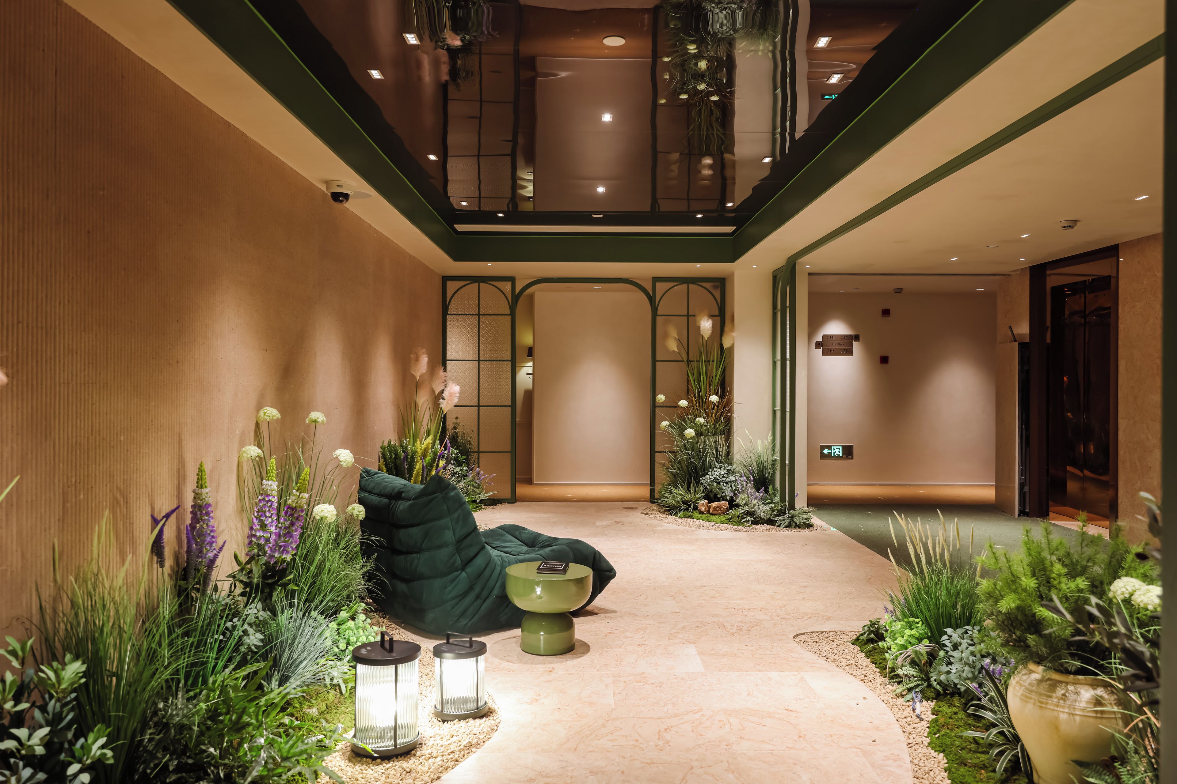 Un hall de hotel con un sofá verde, plantas decorativas y una pequeña mesa con dos faroles.