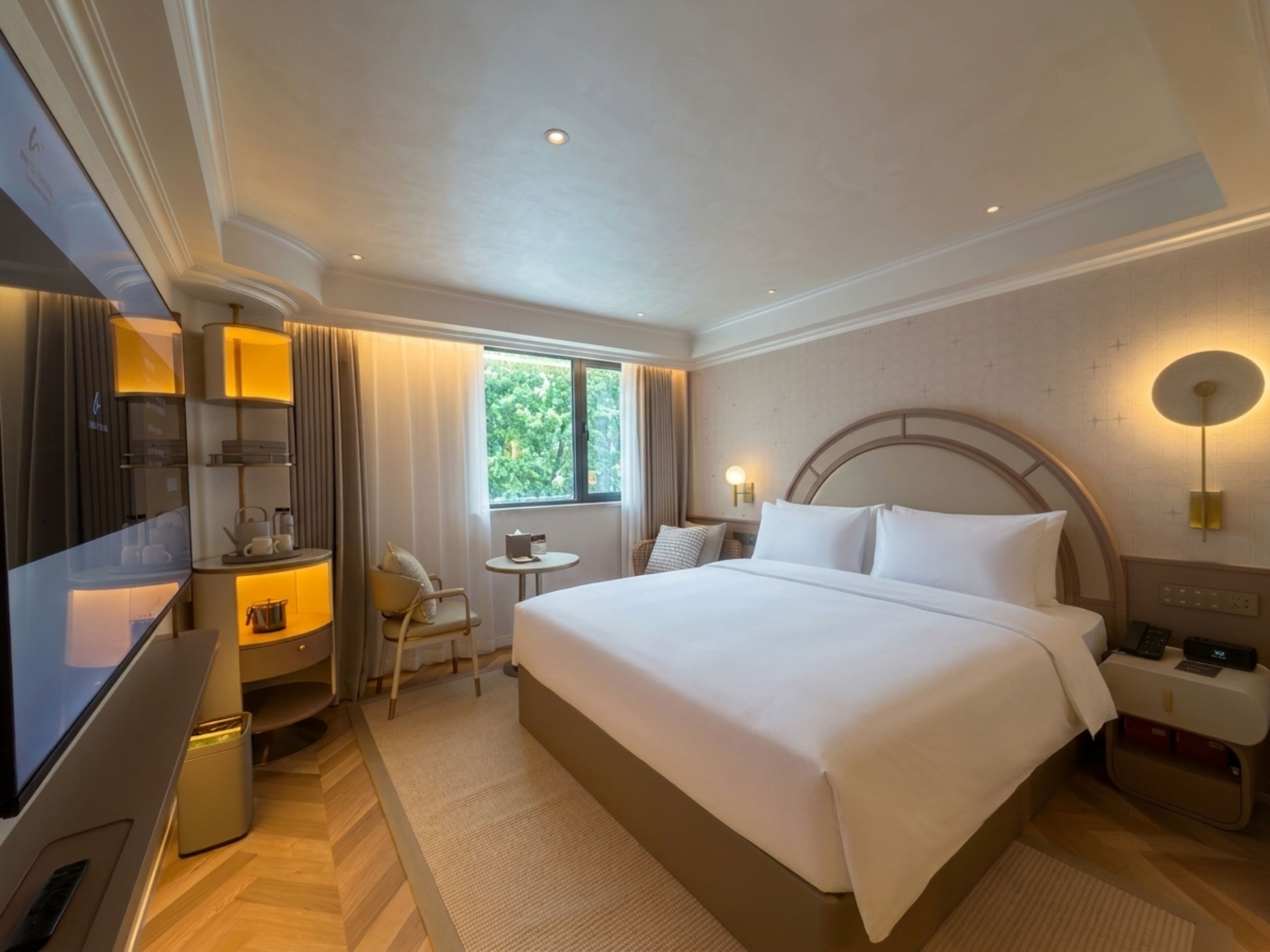 Una habitación de hotel con una cama grande, mesitas de noche, un televisor, una pequeña mesa y una ventana con cortinas.