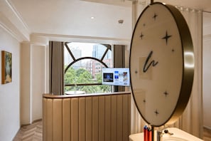 Une chambre d’hôtel avec une grande fenêtre, un miroir rond, une télévision et une vue sur des immeubles et des arbres.