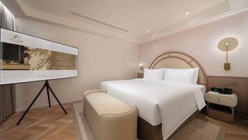 Habitación de hotel con una cama grande, mesitas de noche, un escritorio con una computadora y una pantalla de proyección mostrando información del hotel.