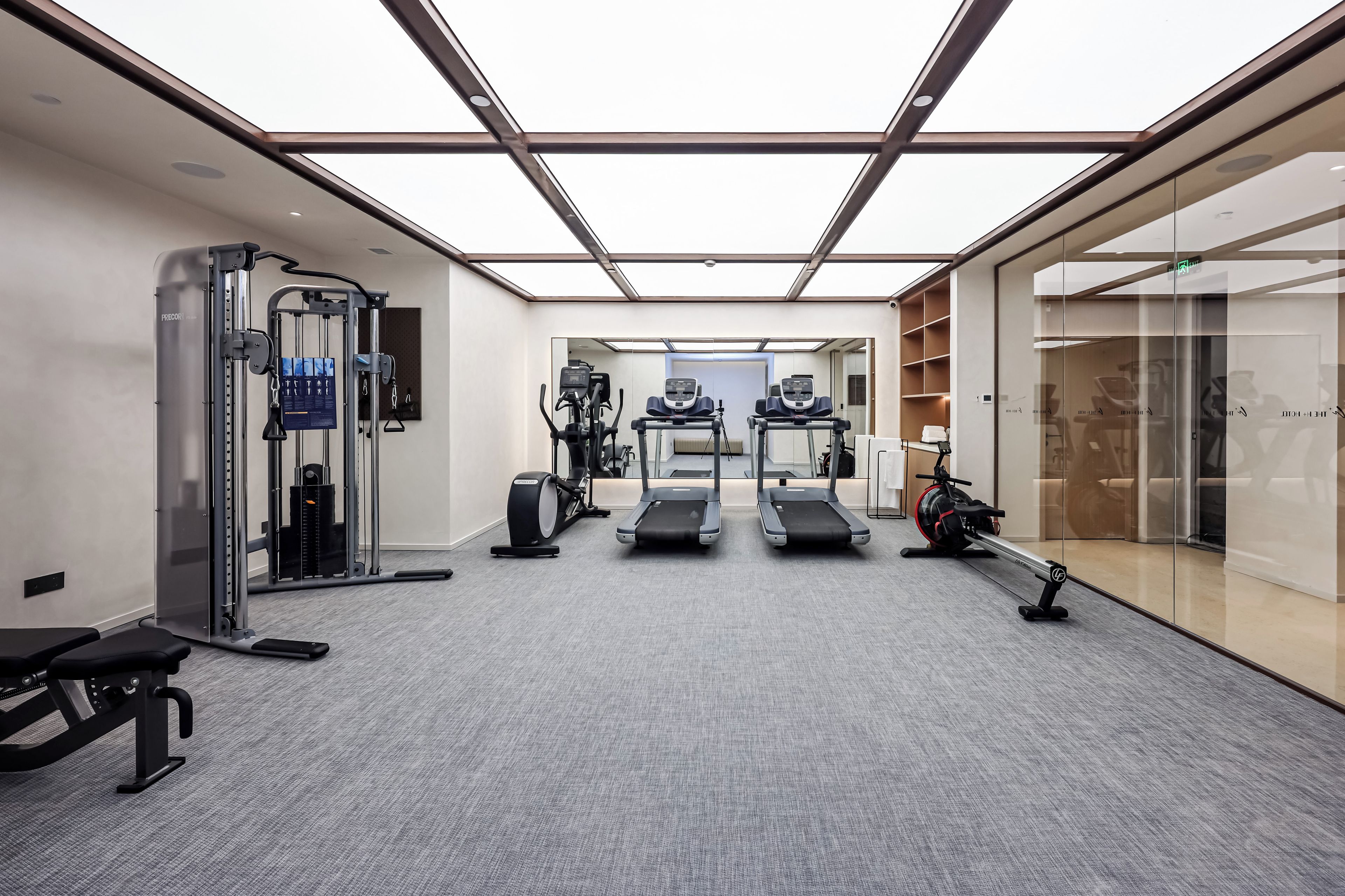 Un gimnasio moderno con equipo de ejercicio, un muro espejado y una puerta de cristal que da a un pasillo.