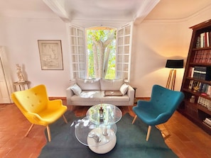 Ein Zimmer mit einer Couch, zwei Sesseln, einem Glastisch, einem Bücherregal und einem Fenster mit Vorhängen.