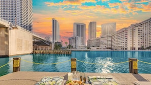 A waterfront view with high-rise buildings in the background, a dock with caution signs, and a table with food and drinks. - 2BR BW Resort - Bay View Fitness Center (Hallandale Beach)