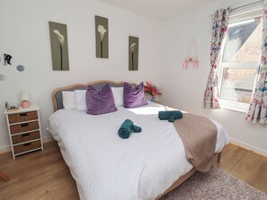 A neatly made bed with a wooden headboard, white bedding, and purple pillows. A small bedside table with a lamp and a book is visible. - The Old Bank House (Morpeth)