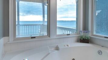 A bathroom with a large bathtub, a window with a view of the sea, and a small plant on the windowsill.