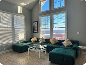 A living room with a sectional sofa, a coffee table, and large windows with blinds. - Modern Soundfront - Sleeps 12 (Manteo)